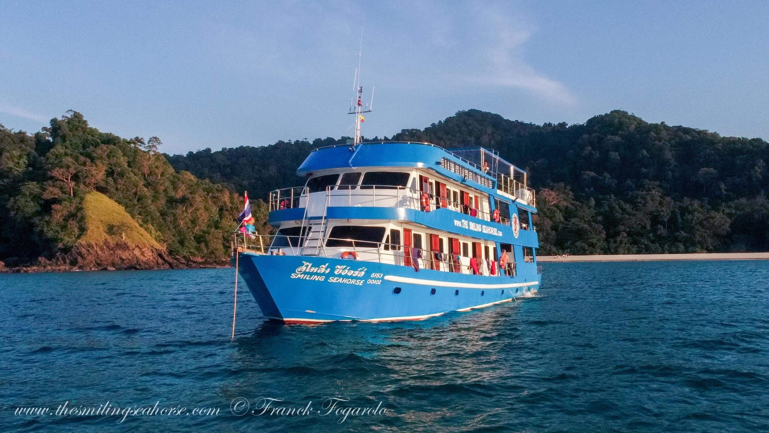 The MV Smiling Seahorse liveaboard dive boat anchored in the calm ocean near a tropical island with green hills and a sandy beach.